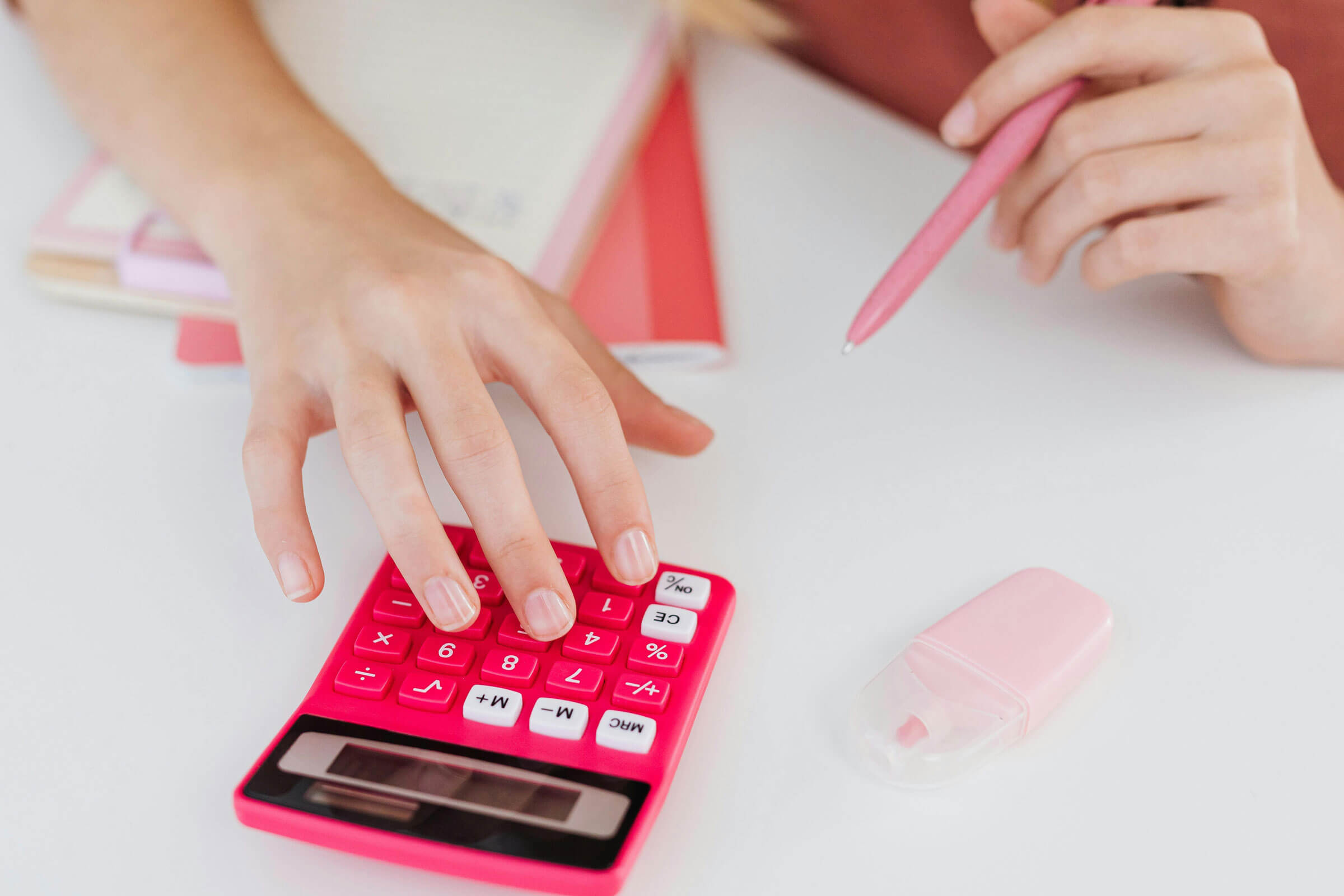 Pair of hands, one had holding a pink pen, the other hand operating a pink calculator.