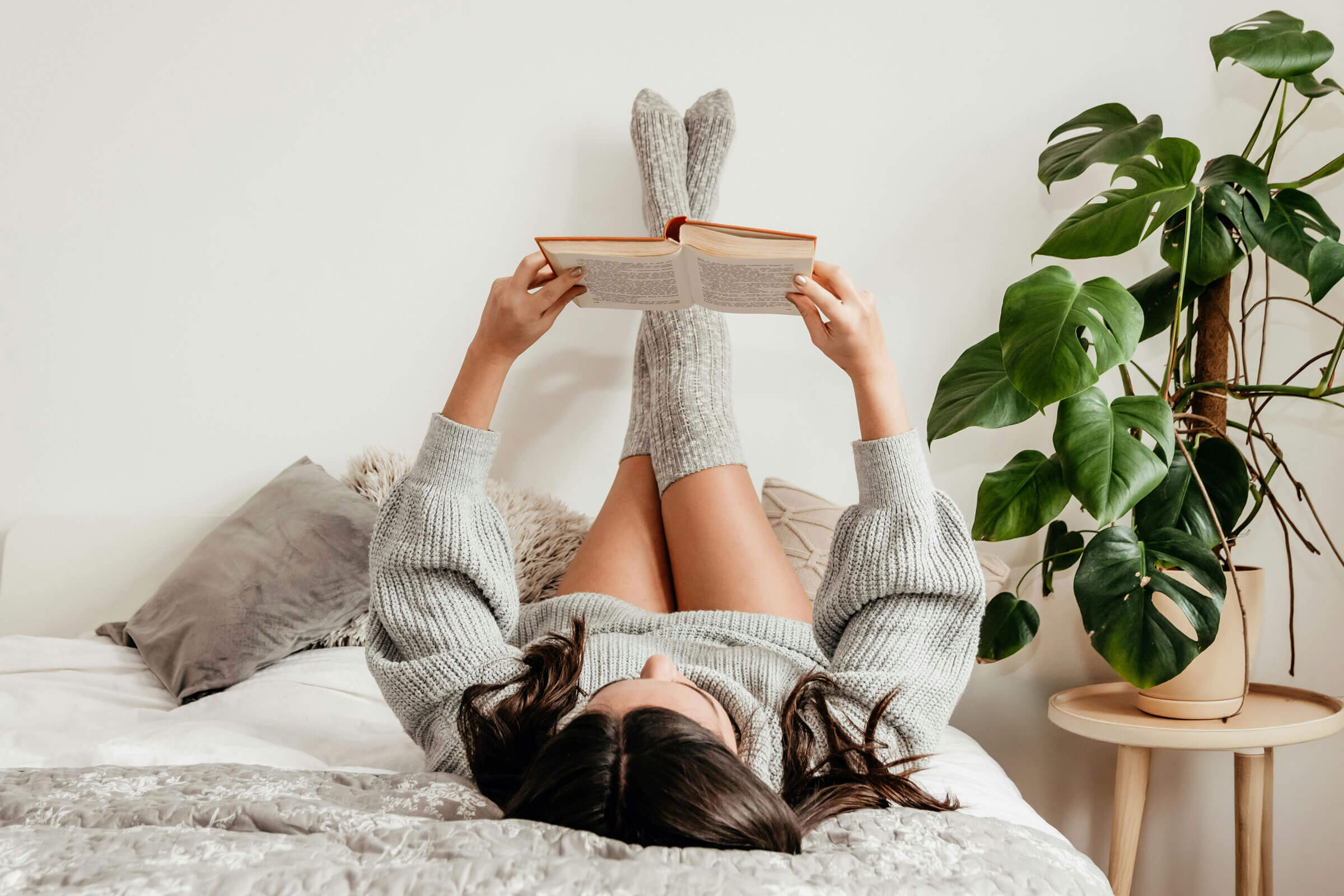 Woman reading book in bed, lying on back with legs leaning against the wall.